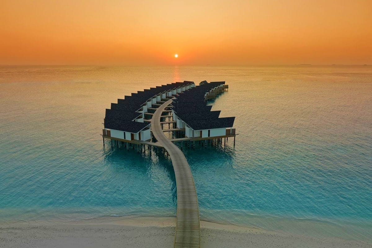 Overwater bungalow resort at sunset with curved wooden boardwalk connecting thatched-roof villas in turquoise tropical waters