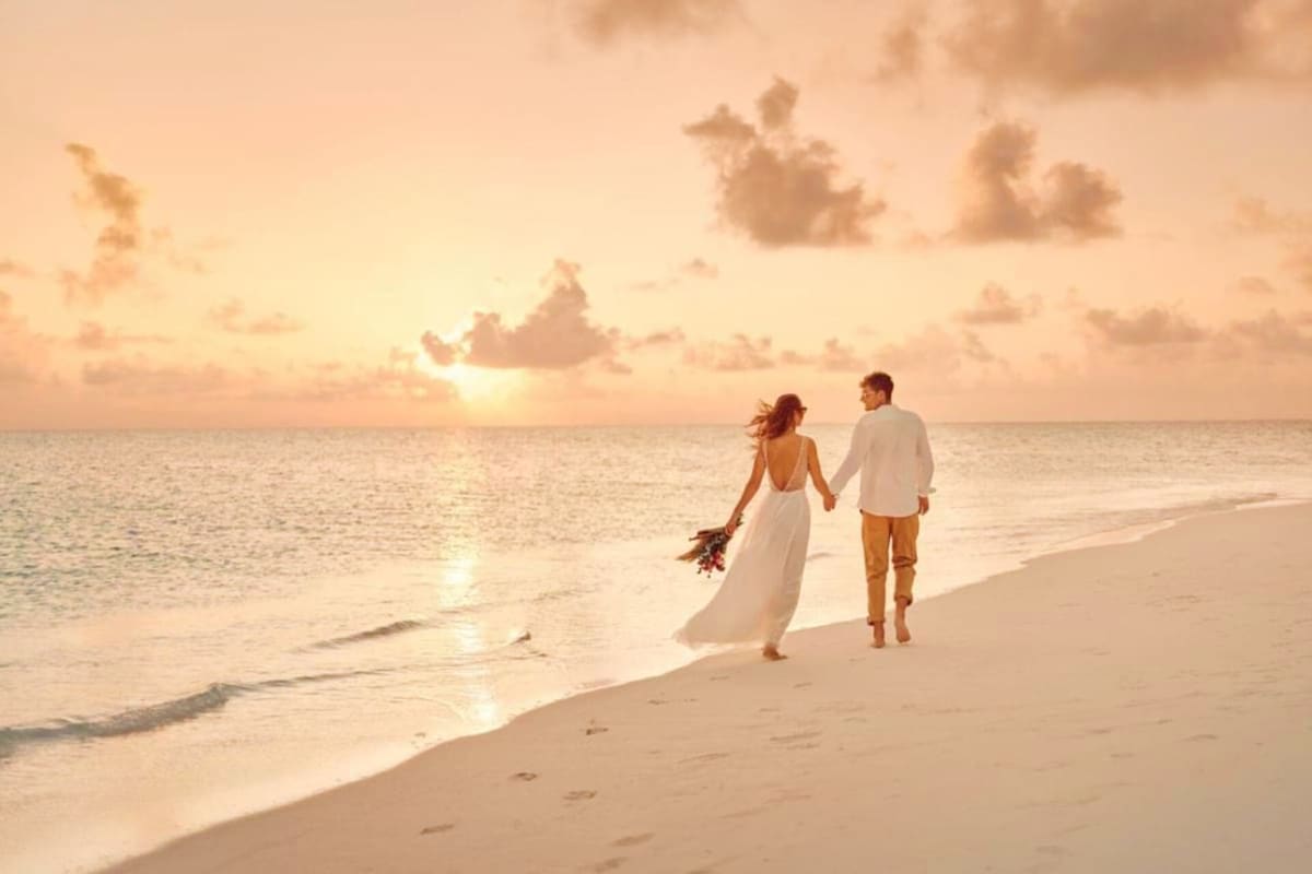 Bride and groom walking hand in hand on a tropical beach at sunset, woman in white wedding dress holding bouquet