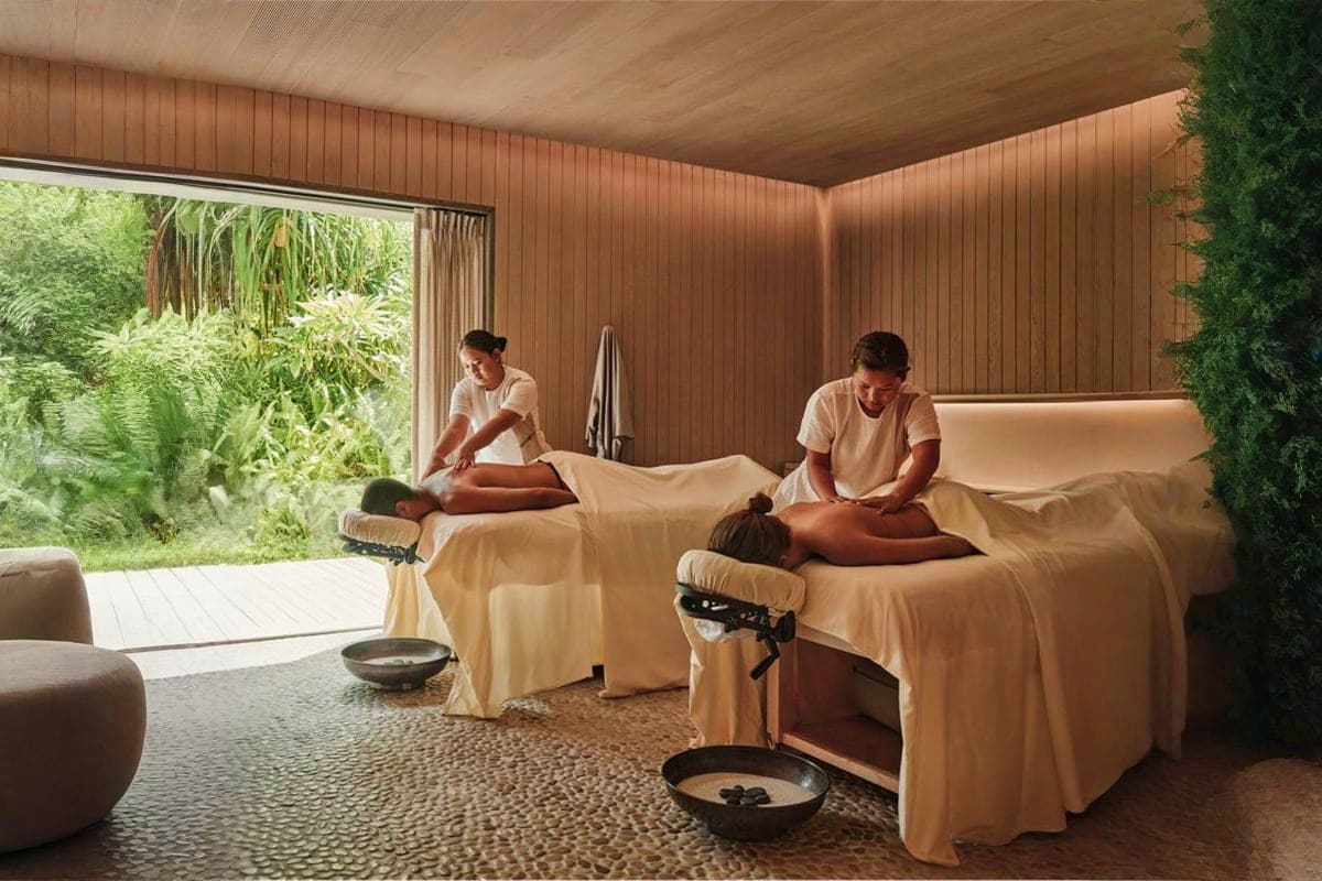 Spa therapists performing massages on two clients in a modern wooden wellness room with garden views and indoor plants.