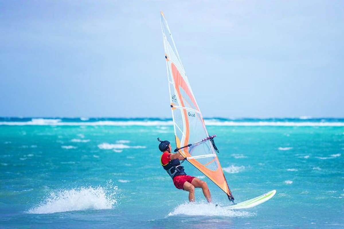 Windsurfer in red and black gear riding waves on turquoise ocean water under clear blue sky