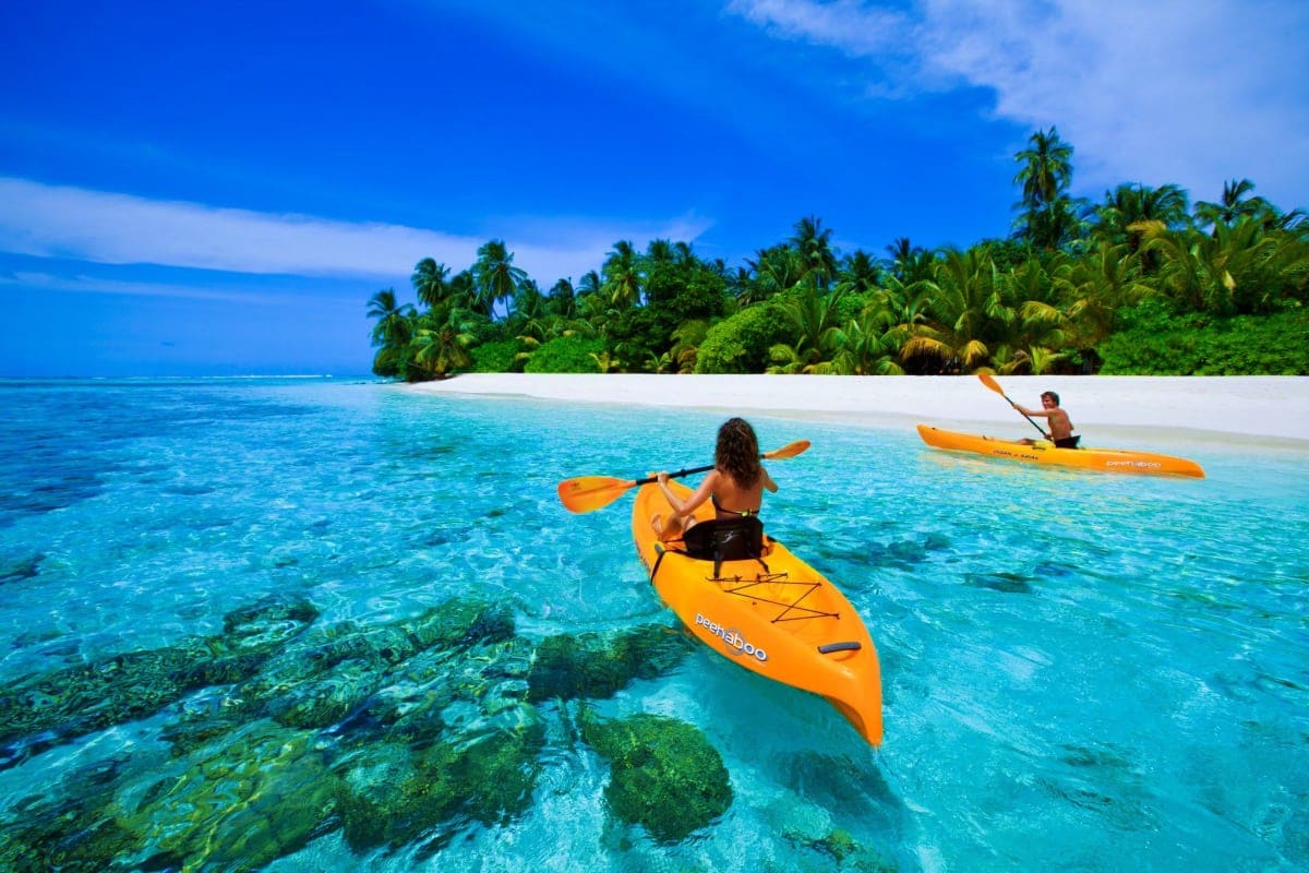 People kayaking in turquoise water near a tropical island with white sand beach and palm trees in the Maldives