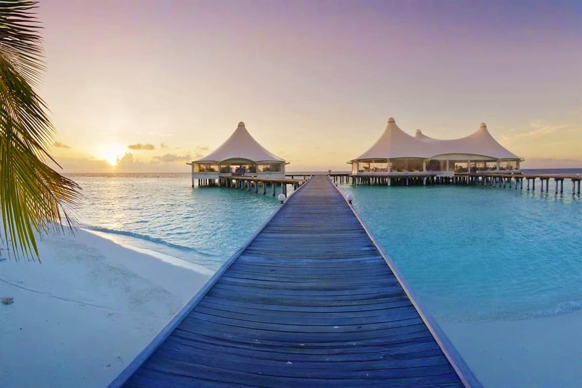 Wooden boardwalk leading to white tent-roofed overwater bungalows at sunrise in turquoise lagoon, Maldives