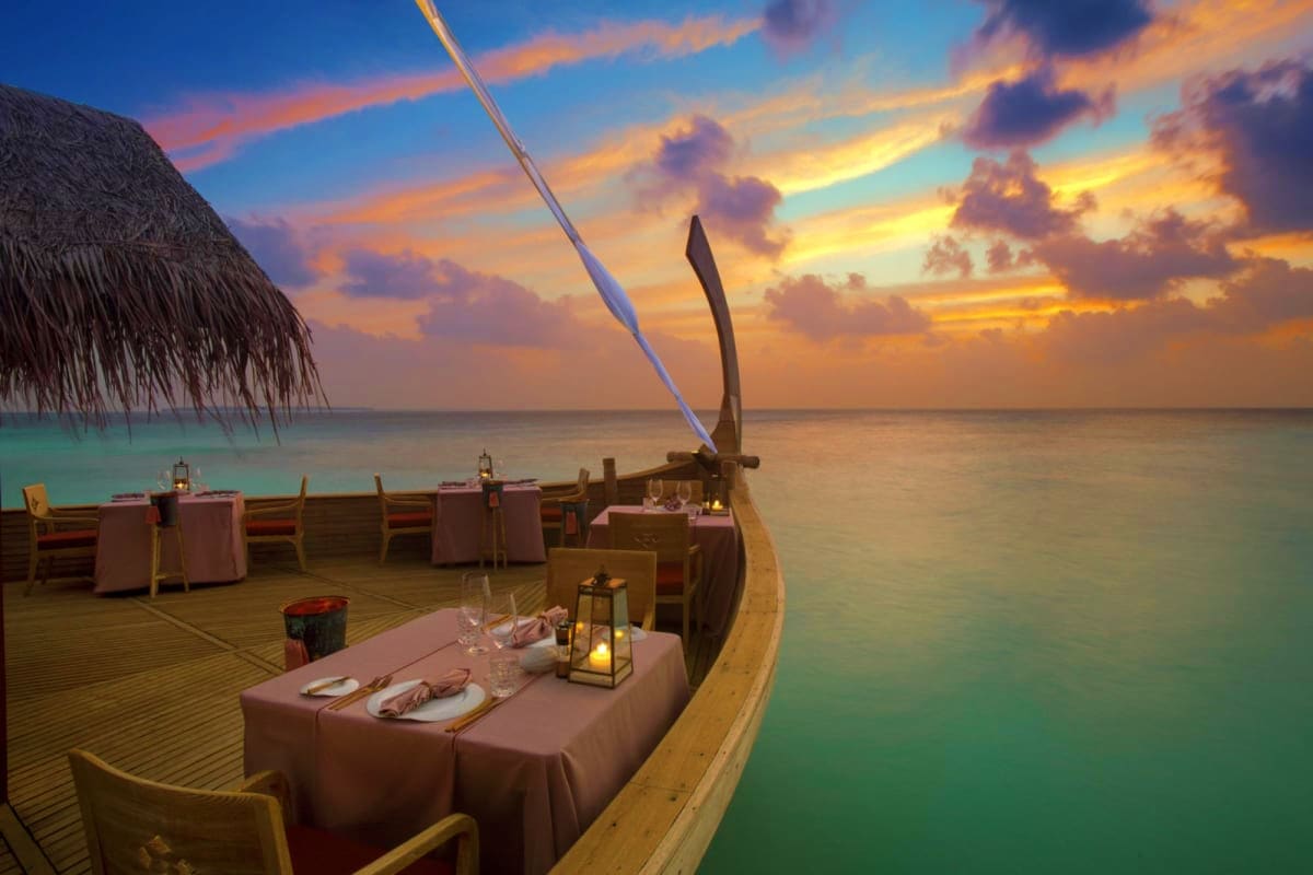 Sunset view of an elegant outdoor dhoni boat dining setup with pink tablecloths, lanterns, and turquoise ocean waters at JW M