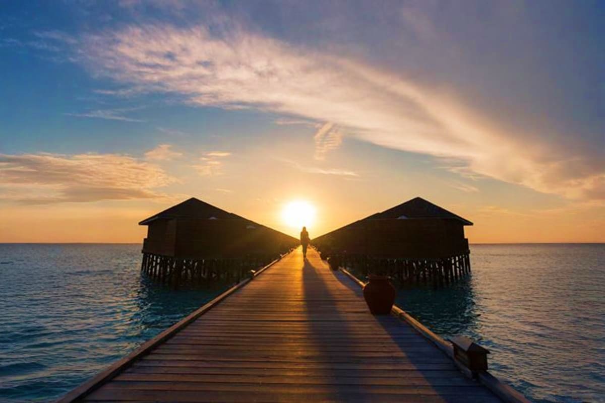 Wooden boardwalk stretching toward sunset over ocean with two thatched-roof beach bungalows on either side at JW Marriott Mal