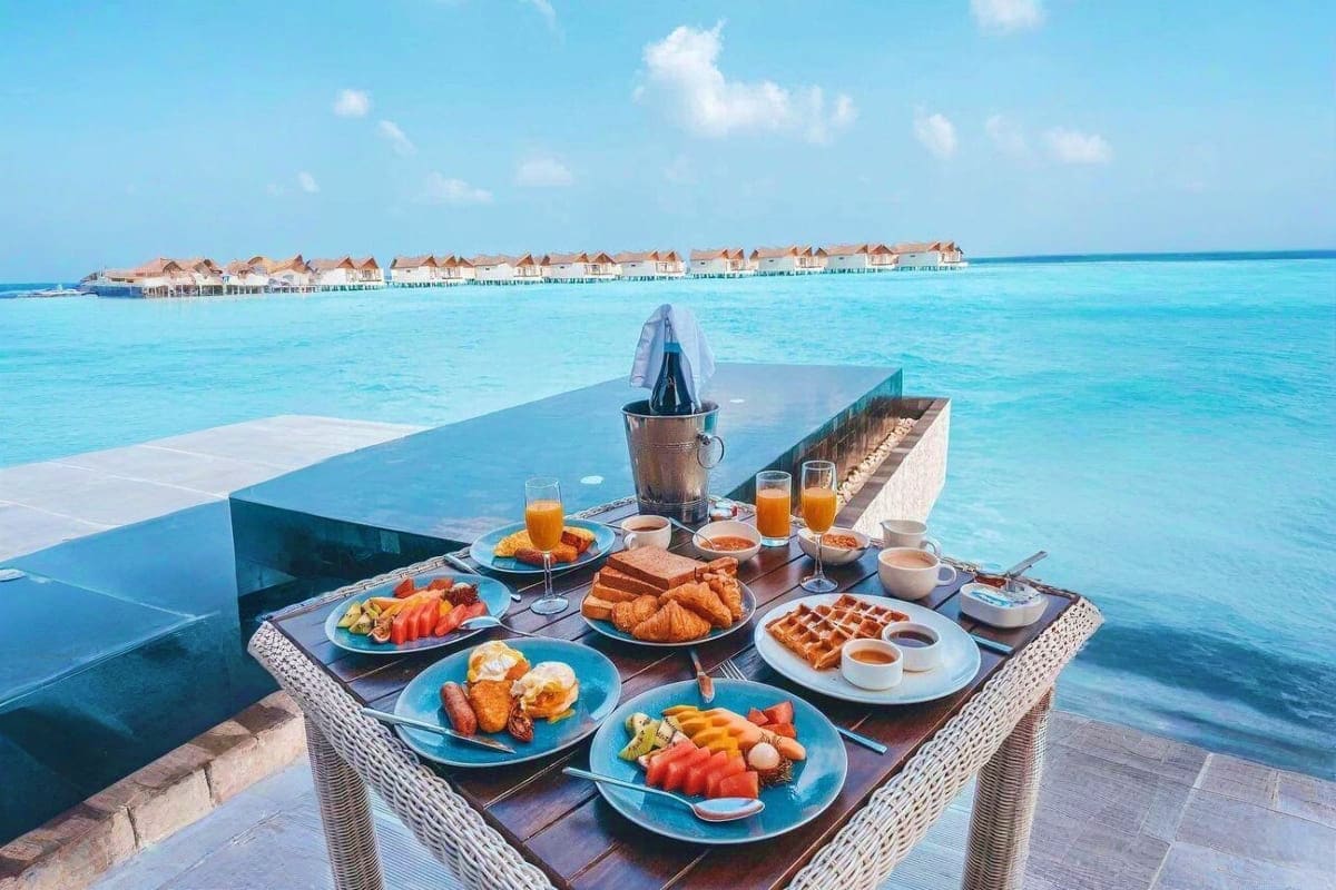 Luxury breakfast spread on overwater deck at JW Marriott Maldives with thatched bungalows and turquoise lagoon in background