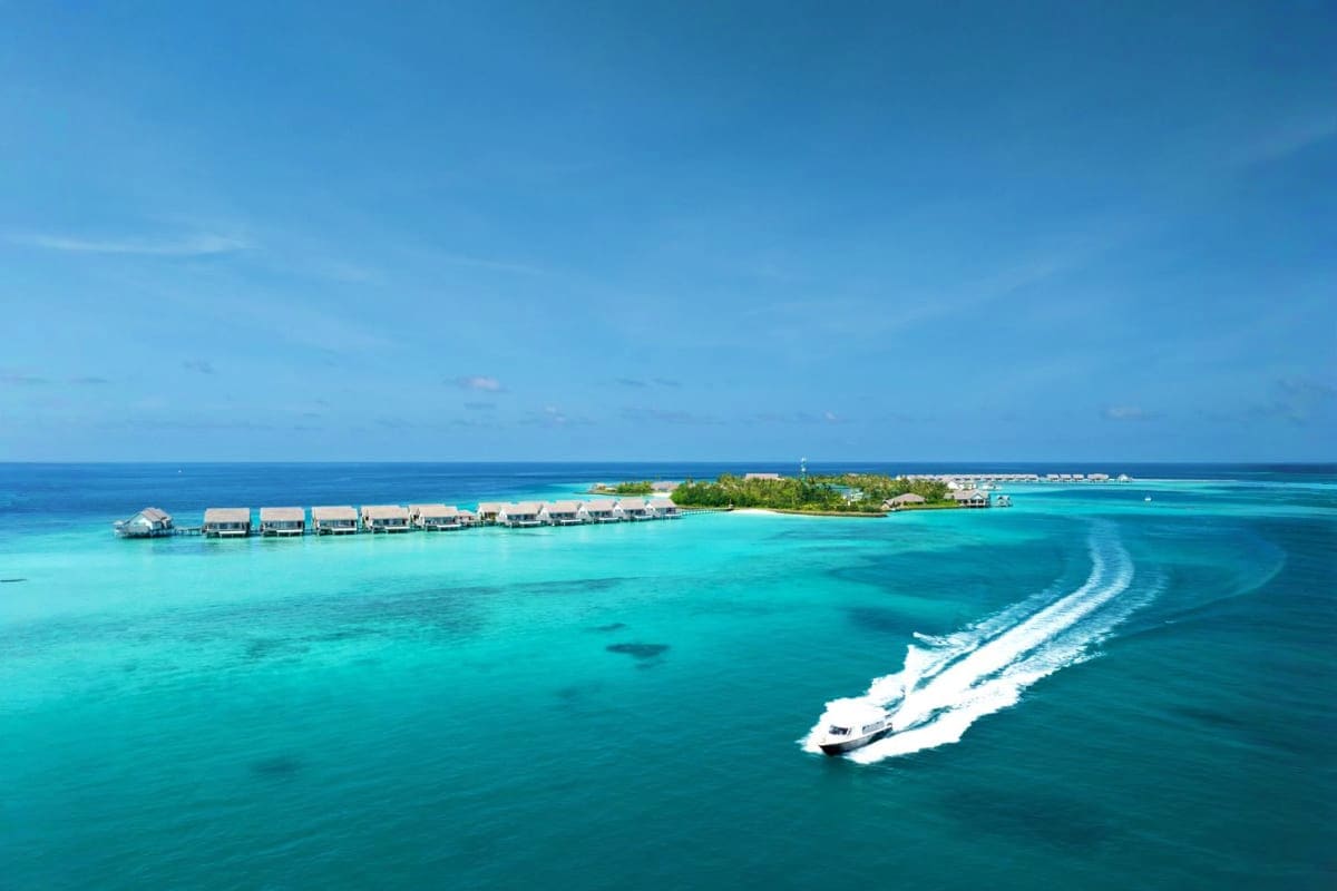 Aerial view of JW Marriott Maldives Resort with overwater bungalows, tropical island, turquoise lagoon, and speedboat in crys