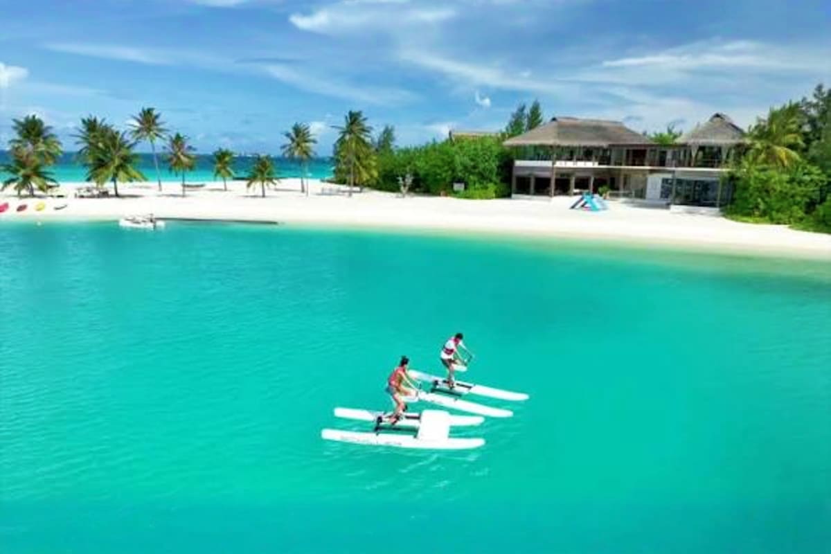 Three people paddling traditional Maldivian outrigger canoes in turquoise water with white sand beach and resort buildings wi