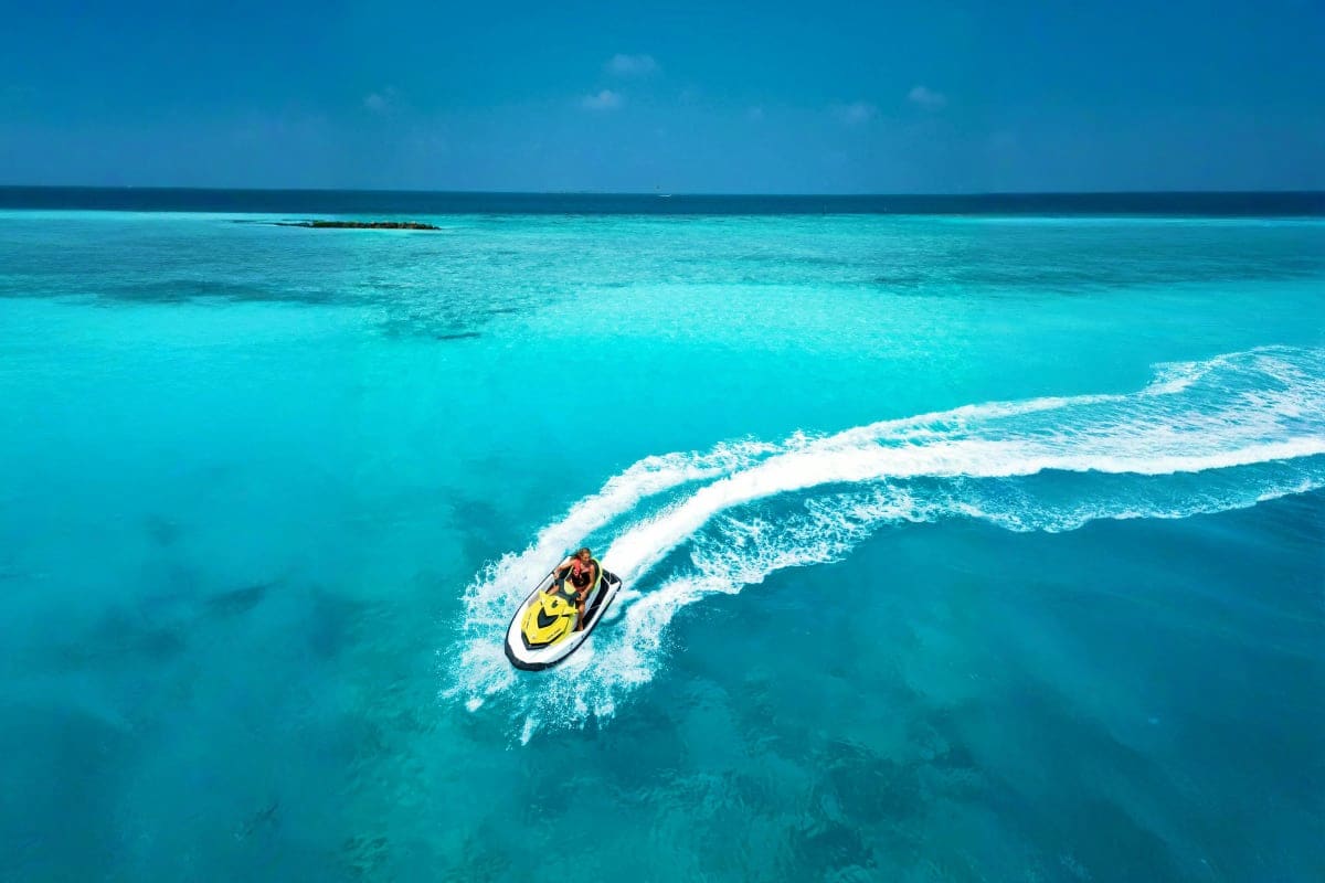 Aerial view of a yellow and white jet ski with rider creating white wake waves in turquoise ocean waters near Sun Siyam Olhuv