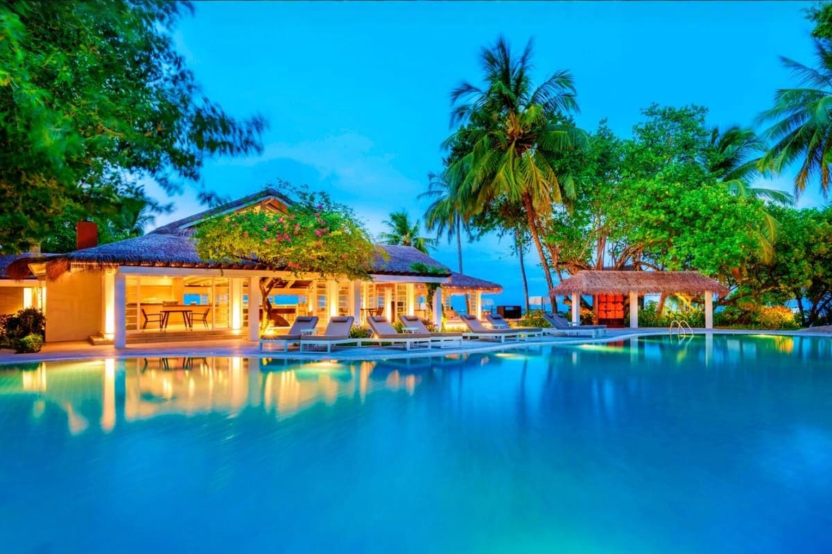 Illuminated beachfront resort with pool, thatched-roof bungalows, palm trees, and twilight sky at Sheraton Maldives Full Moon