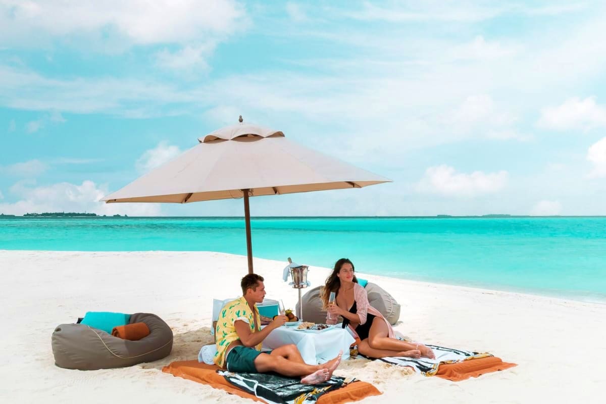 Two people relaxing under a beige umbrella on white sand beach with turquoise water and blue sky in background