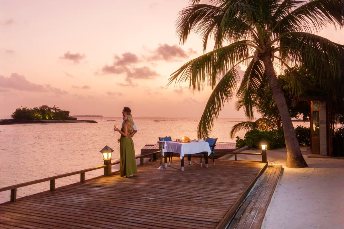 Woman in green dress standing on wooden dock at sunset with dinner table set up overlooking calm ocean and palm trees at trop
