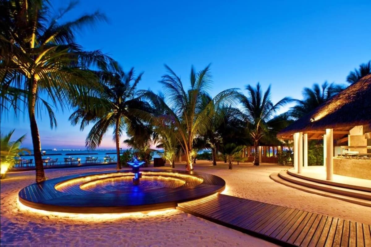 Illuminated circular fountain with blue water feature on sandy beachfront at dusk, flanked by palm trees and modern resort ar