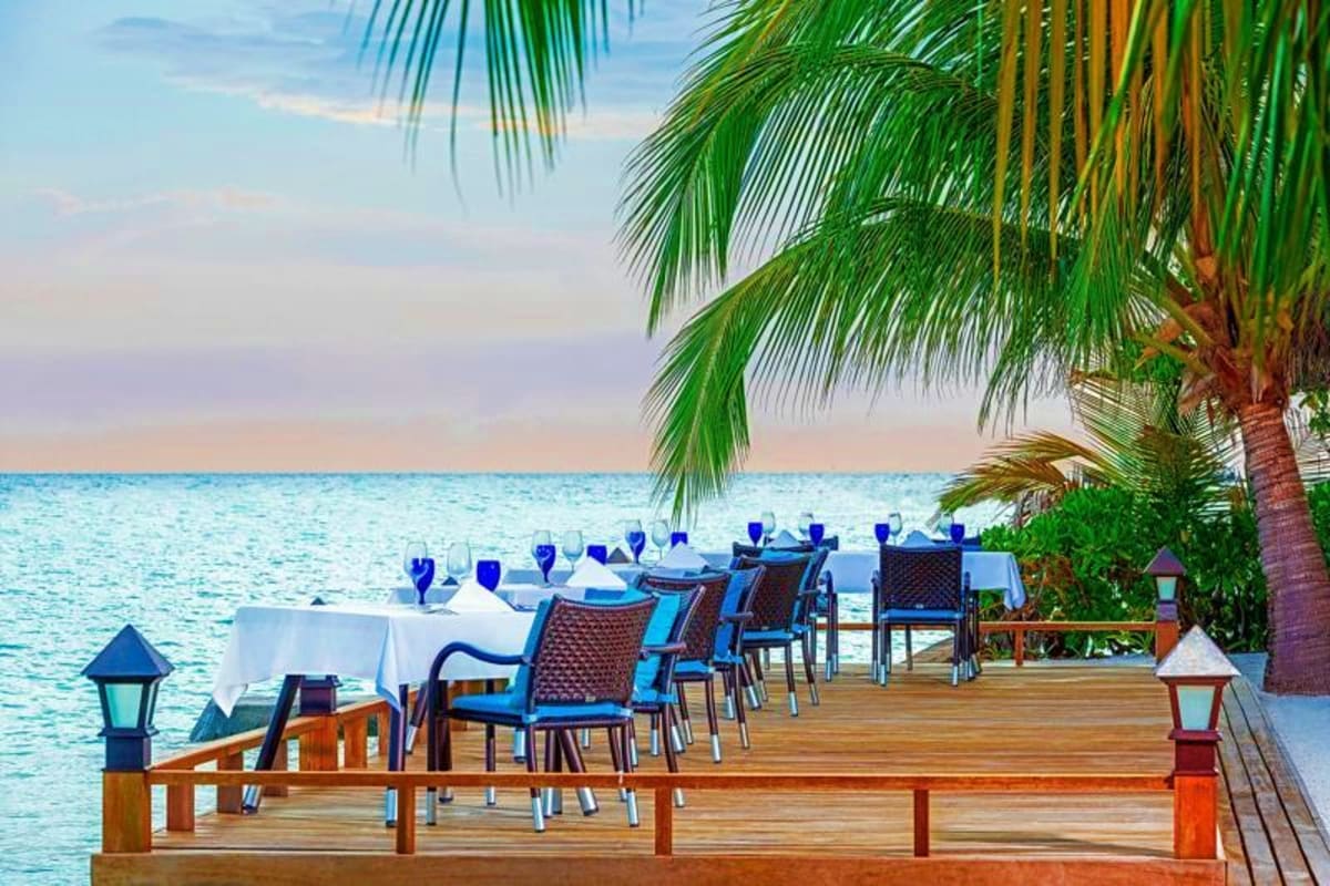 Oceanfront wooden deck dining setup with blue chairs and white tablecloth, palm trees, and turquoise sea at Sheraton Maldives