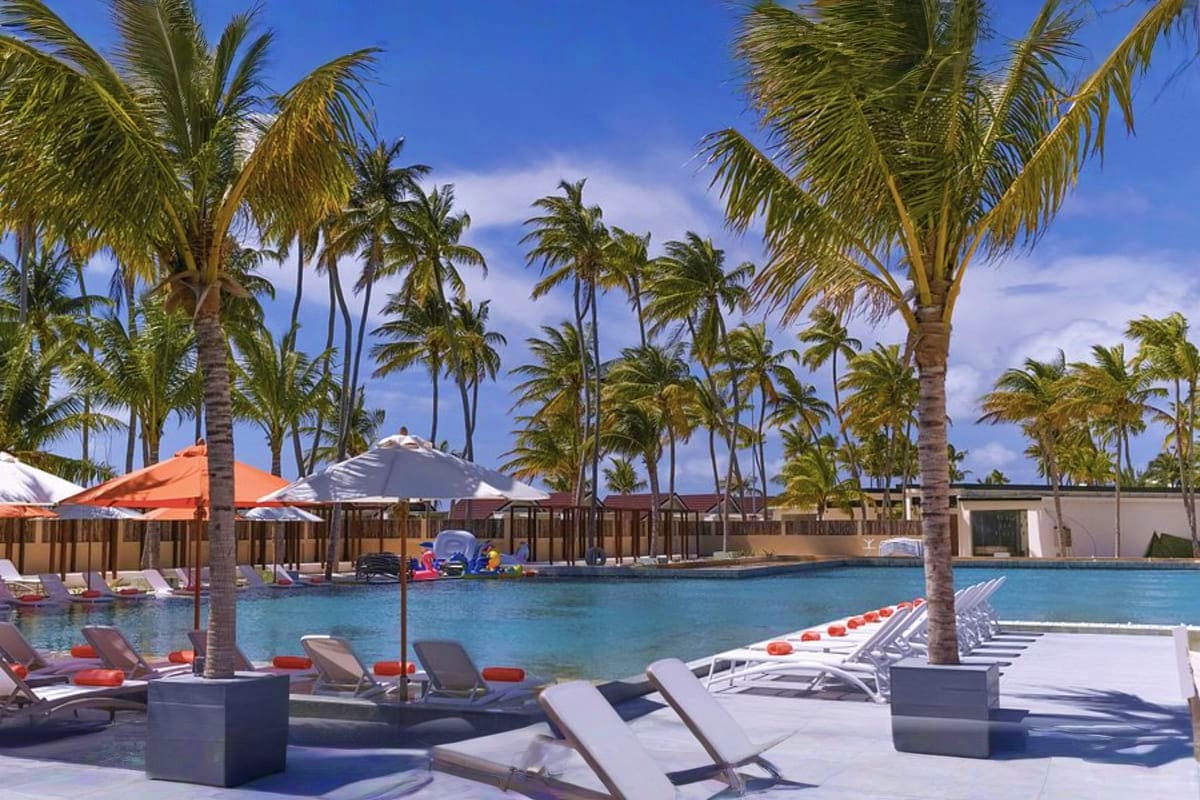 Tropical resort pool area with palm trees, lounge chairs, orange and gray umbrellas, and calm blue water under clear sky at O
