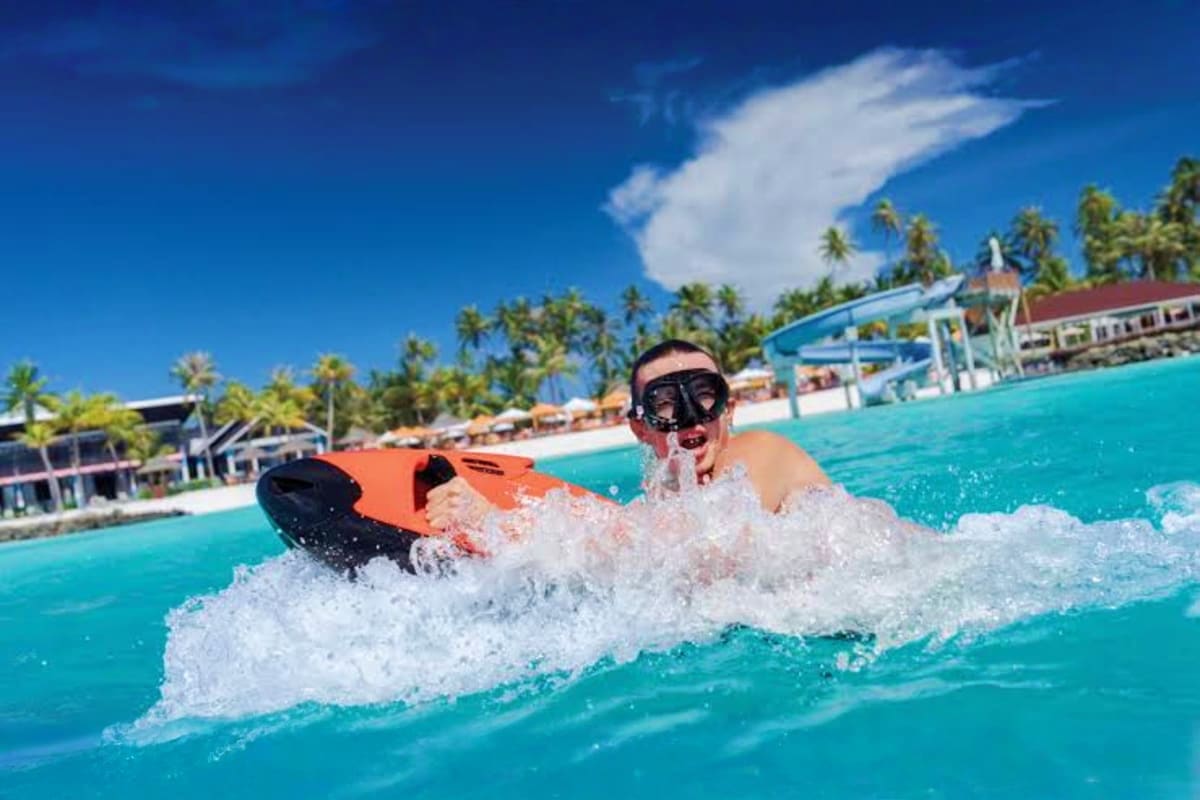Child wearing snorkel gear swimming with orange kickboard in turquoise lagoon at tropical resort with water slide and palm tr