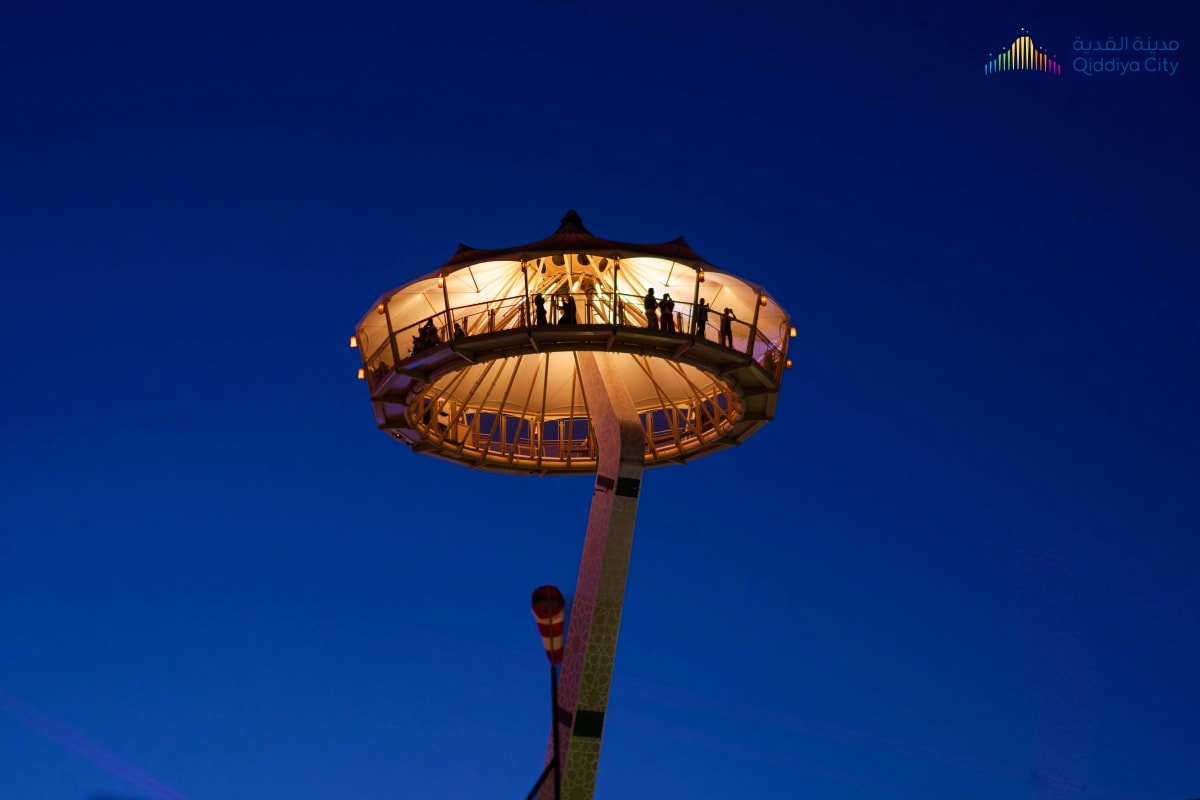 Illuminated observation tower at twilight with visitors on circular viewing platform against deep blue sky, Qiddiya City