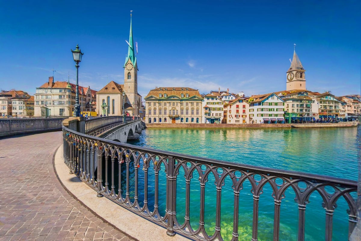 Swiss Italian Explorer Zurich waterfront promenade with ornate railing overlooking turquoise river, historic church spires, and colorful buildings u