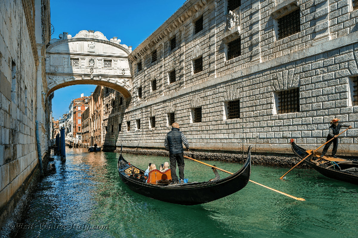Swiss Italian Explorer Gondolier poling traditional black gondola under white Bridge of Sighs in Venice, Italy with historic stone buildings and tur