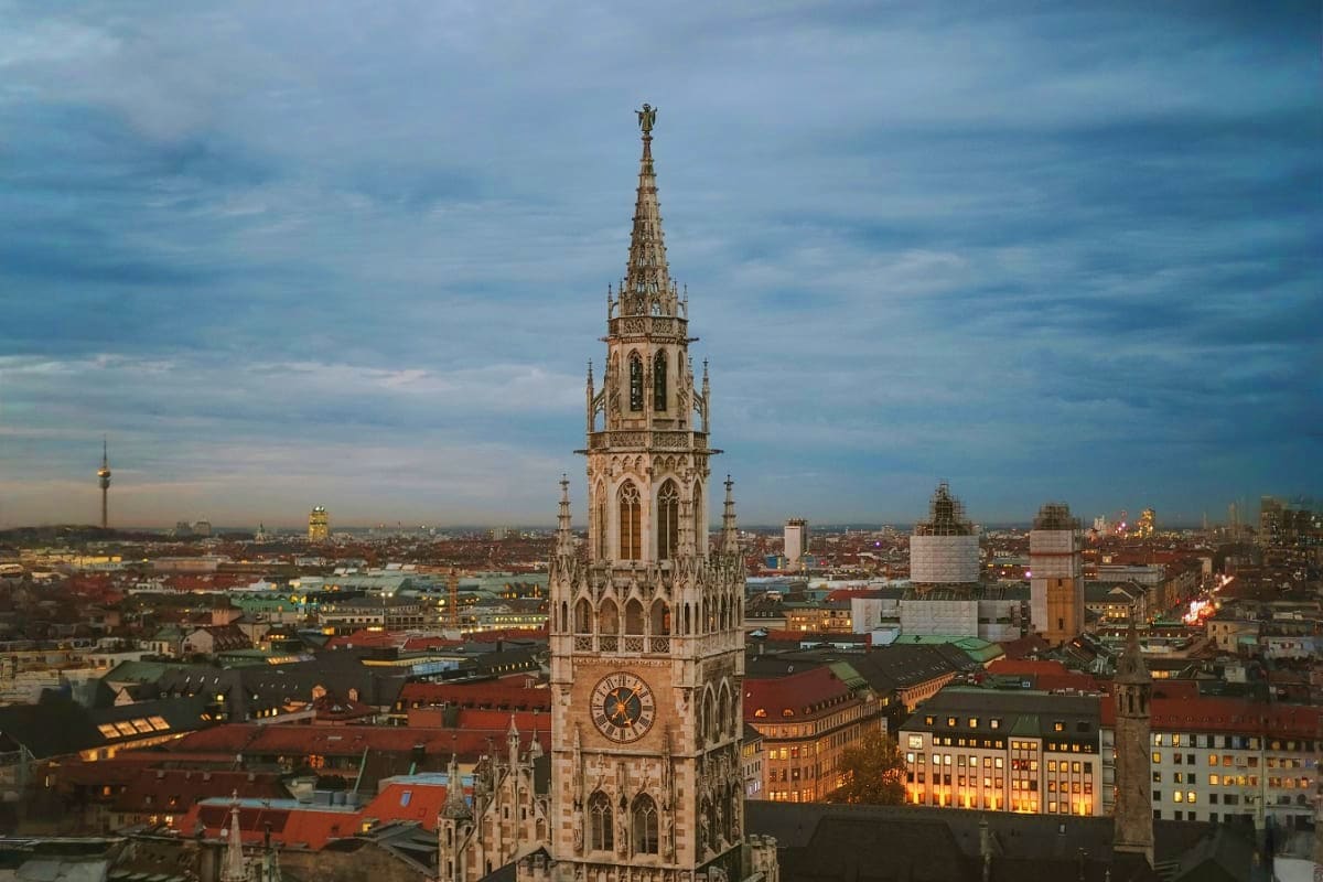 Gothic tower with clock and spire dominating Munich cityscape at dusk with illuminated buildings below