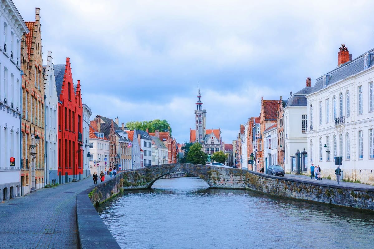 Medieval canal street in Bruges with historic stone bridge, colorful gabled buildings, and church spire in background