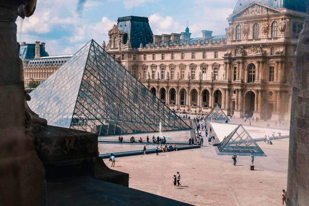 Louvre Museum courtyard in Paris with glass pyramid entrance and classical palace architecture, tourists walking in the plaza