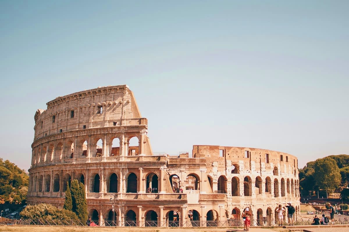 Italy and Switzerland Ancient Roman Colosseum in Rome with partially ruined stone arches and multiple tiers under clear sky, tourists walking nearb