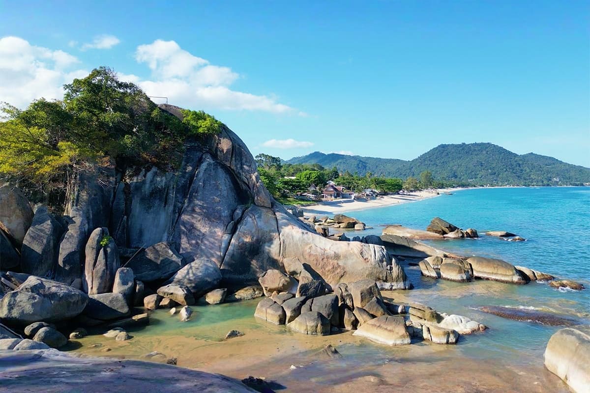 Classic Koh Samui Rocky beach cove with large granite boulders, turquoise water, sandy shore, forested hills, and beachfront buildings in Koh S