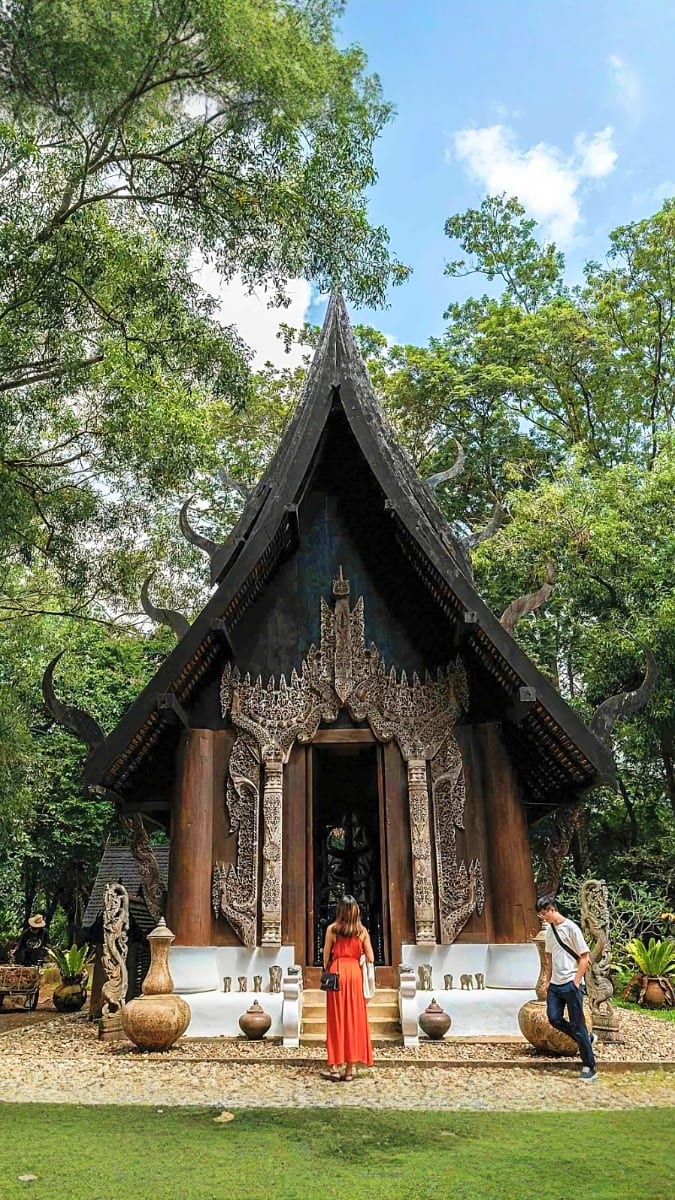 Traditional Thai Buddhist temple with black peaked roof and ornate wooden carvings surrounded by lush green trees, with visit