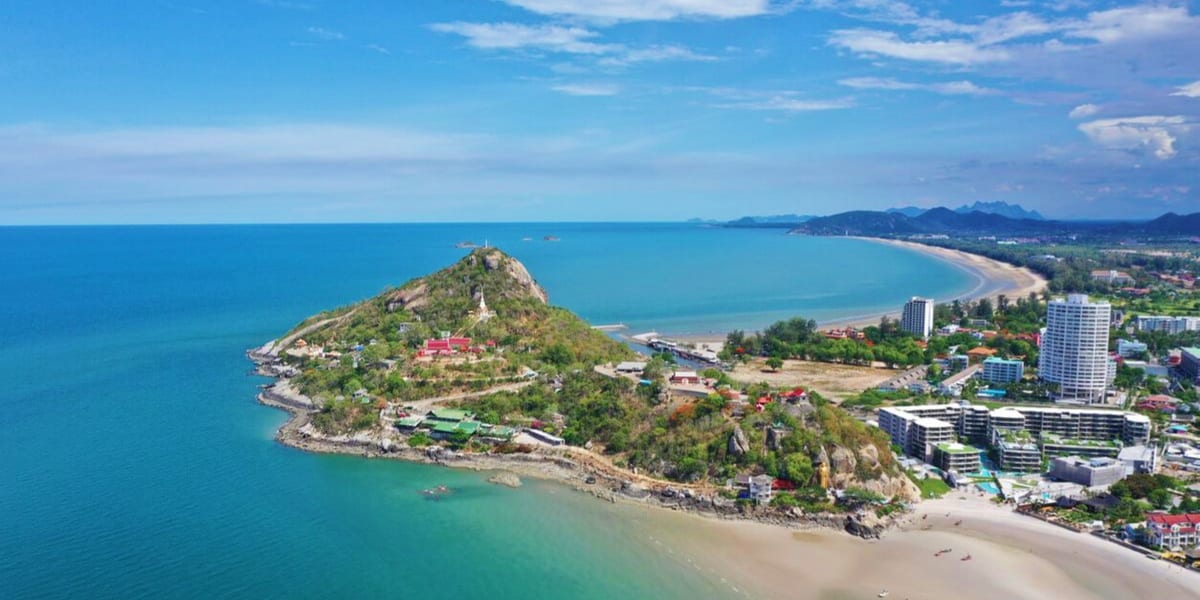Aerial view of Hua Hin headland with golden beach, turquoise sea, temple on rocky peninsula, and coastal resort buildings