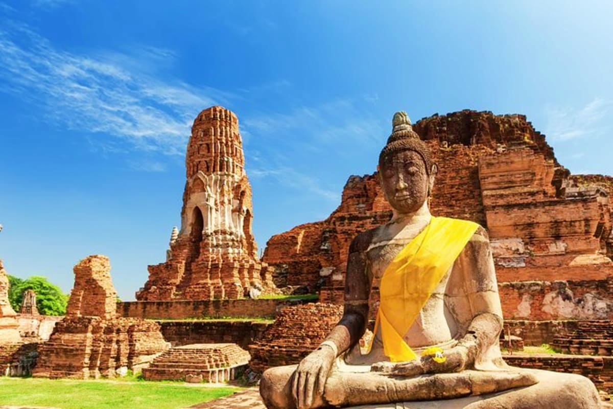 Ayutthaya Full Day Tour Ancient Buddha statue in yellow robe at Ayutthaya temple ruins with ornate brick tower and historical structures under blue s