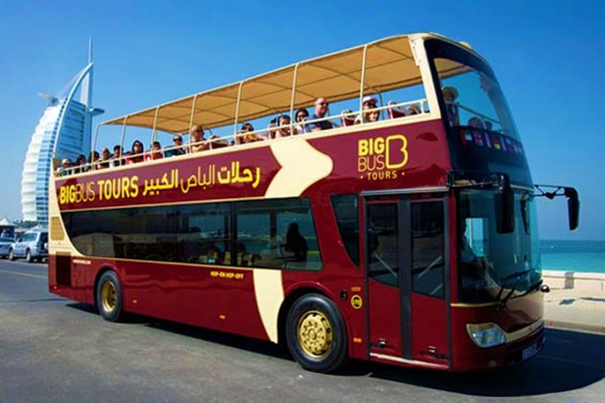 Red and cream double-decker tour bus with open upper deck and passengers, Burj Al Arab hotel visible in background, Dubai bea