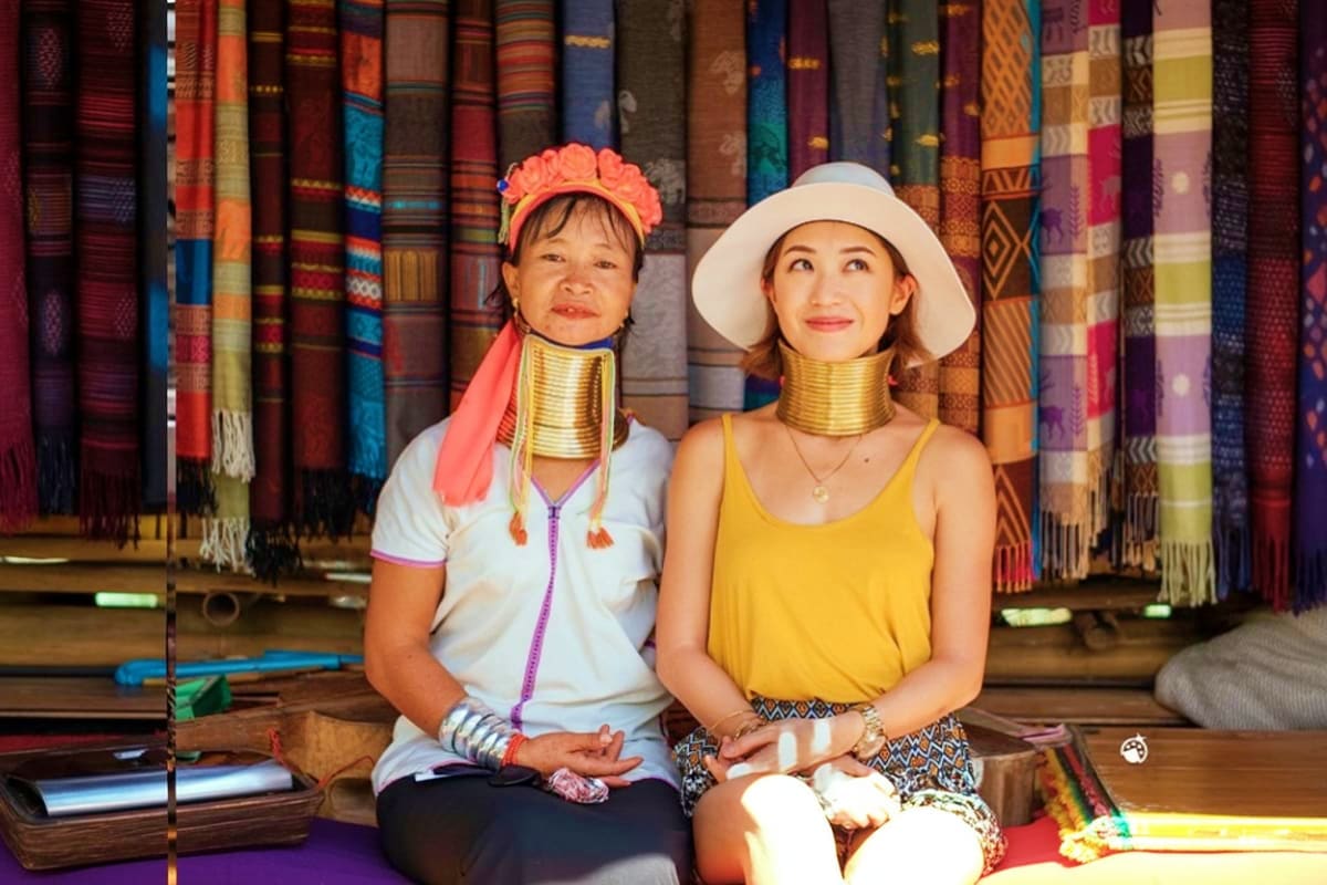 Chiang Rai City Tour Two women wearing traditional Karen long neck brass coils and traditional clothing sitting in front of colorful woven textile