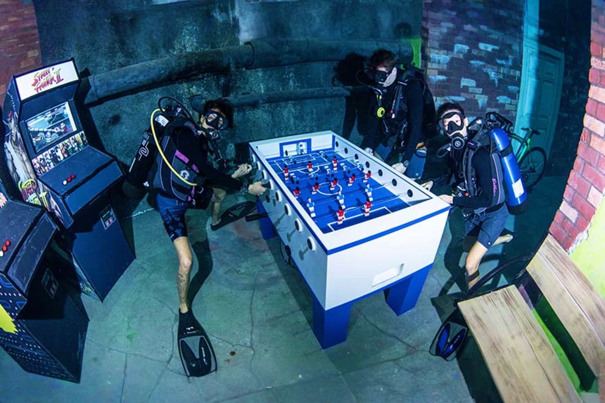 Four scuba divers playing foosball underwater at Deep Dive Dubai, with a Street Fighter II arcade machine visible in the subm