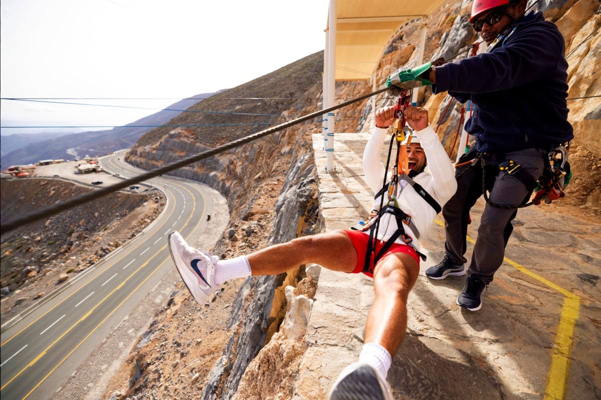 Adventurer ziplining off mountain cliff with safety gear, scenic winding mountain road visible far below in desert landscape