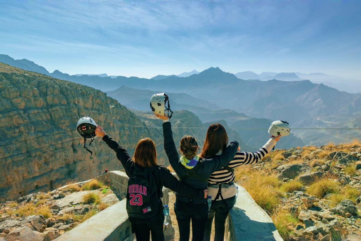 Jebel Jais Zipline Three women celebrating on a mountain ridge holding zipline helmets with dramatic mountain landscape and valleys visible belo