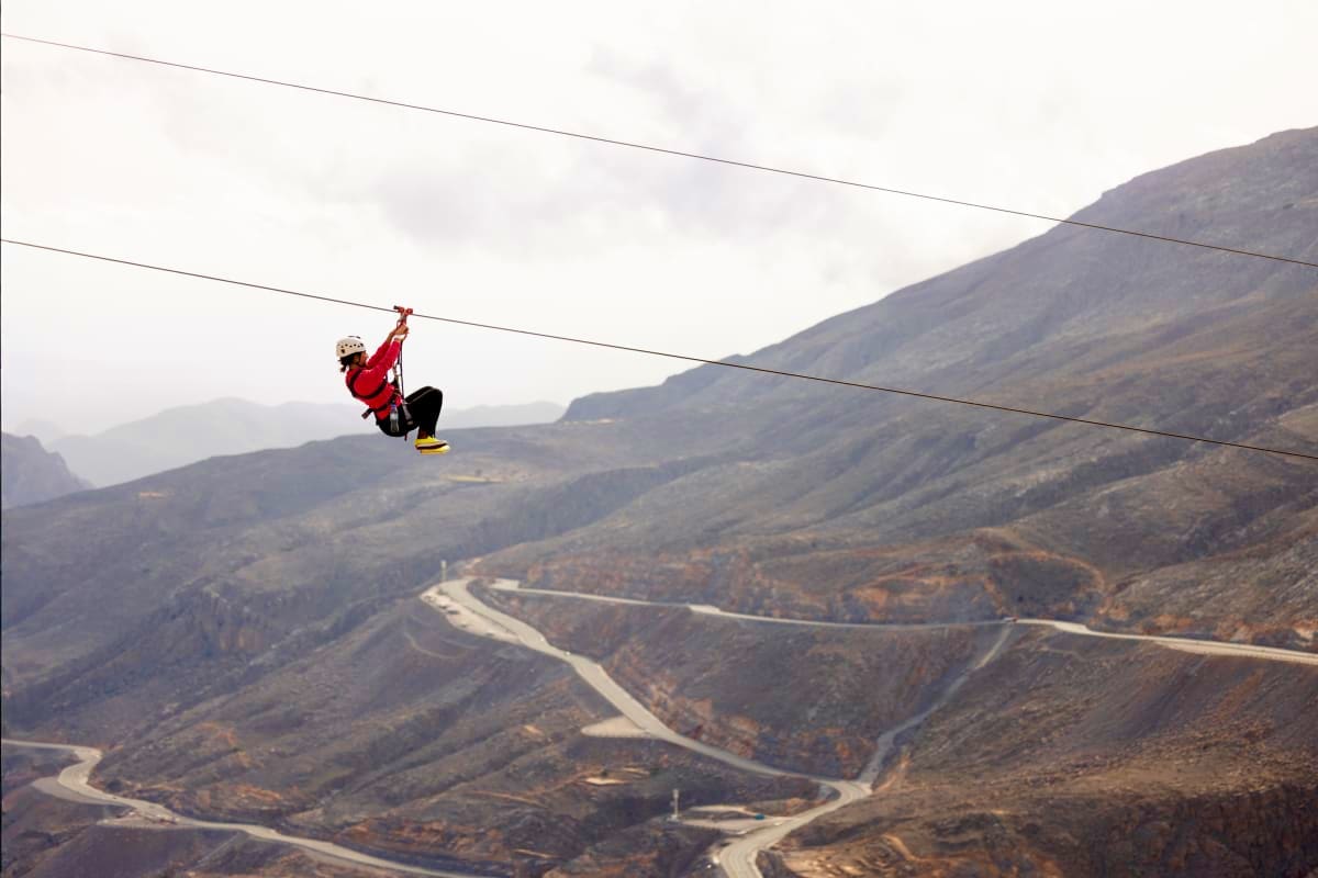 Jebel Jais Zipline Person ziplining on Jebel Jais with mountainous terrain and winding road visible below in United Arab Emirates