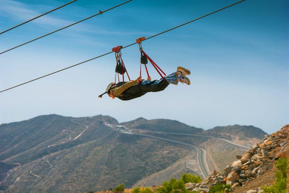 Jebel Jais Zipline Person zip-lining horizontally on cable with mountainous desert landscape and winding roads below in clear blue sky