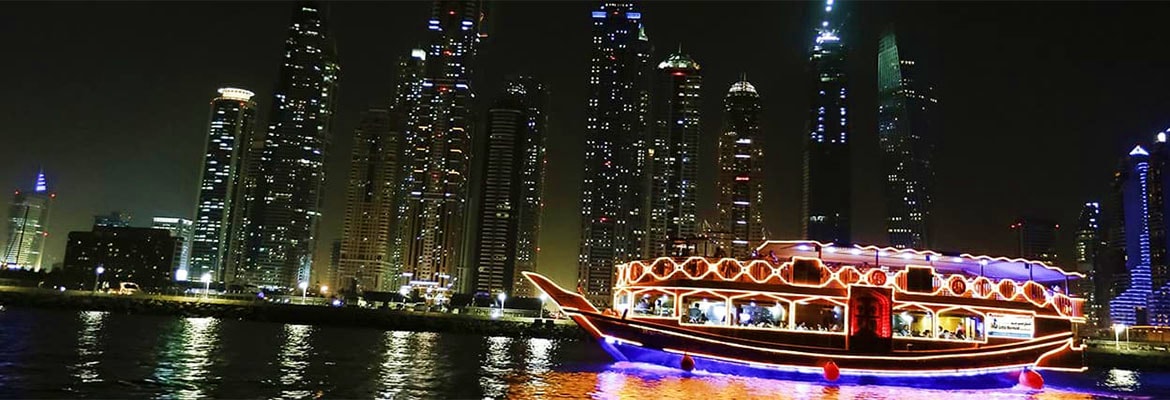 Illuminated traditional Arabian dhow boat with red and gold decorative lighting on marina water at night with downtown city s