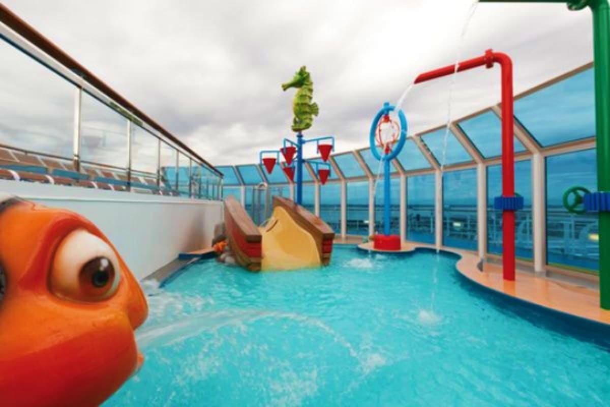 Indoor children's water park on cruise ship with orange fish character, slides, and ocean view through glass ceiling