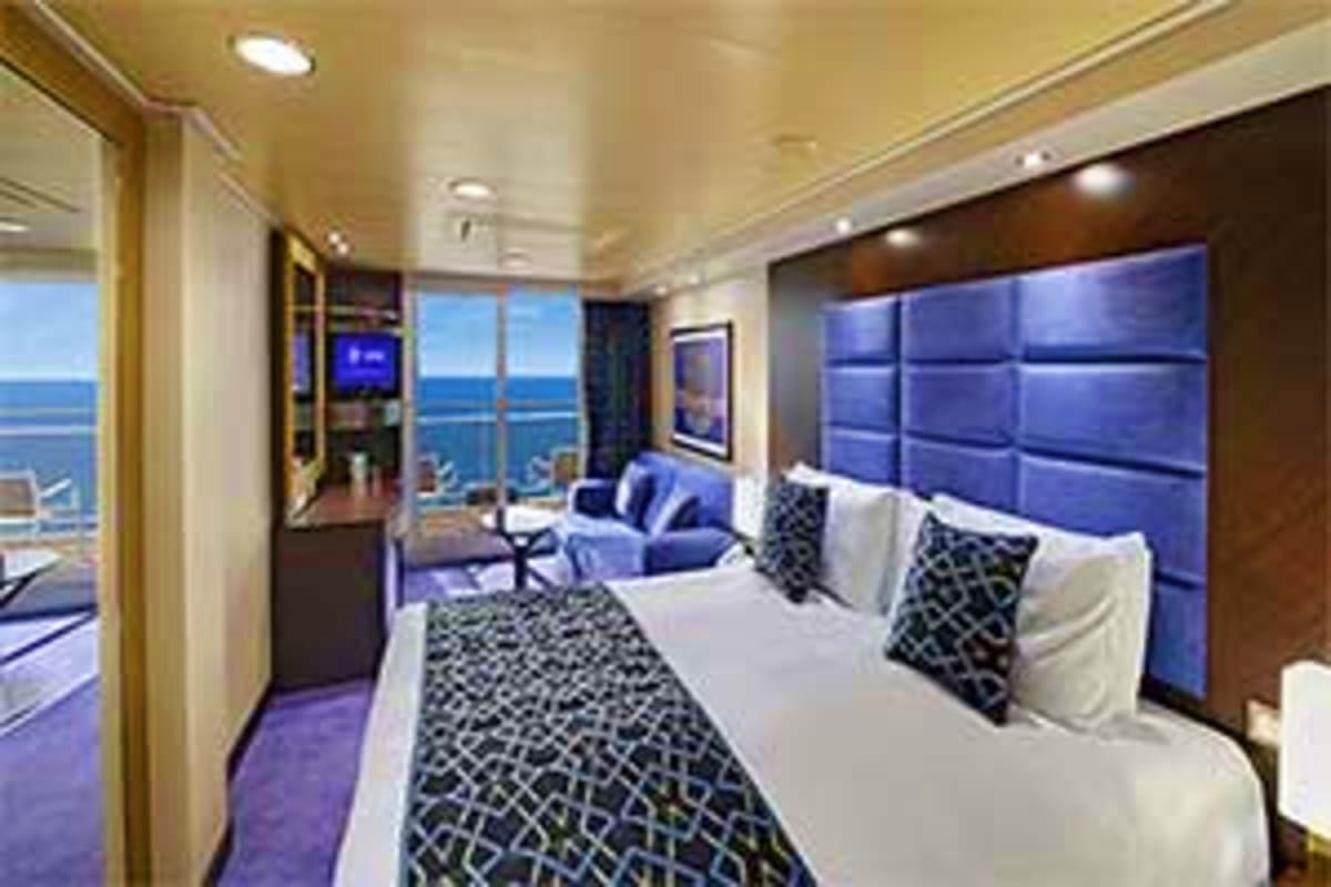 Luxury cruise ship stateroom with ocean views, white bed with blue headboard, seating area, and panoramic windows overlooking