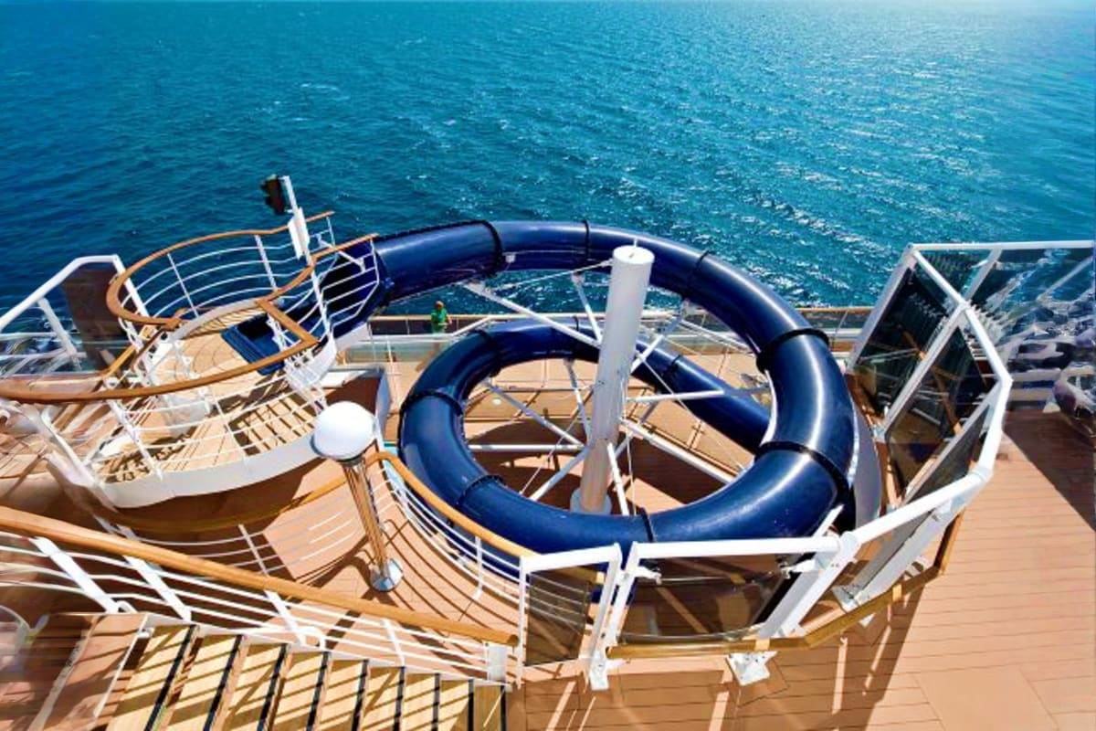 Cruise ship deck with blue waterslide, loungers, and railing overlooking turquoise ocean waters
