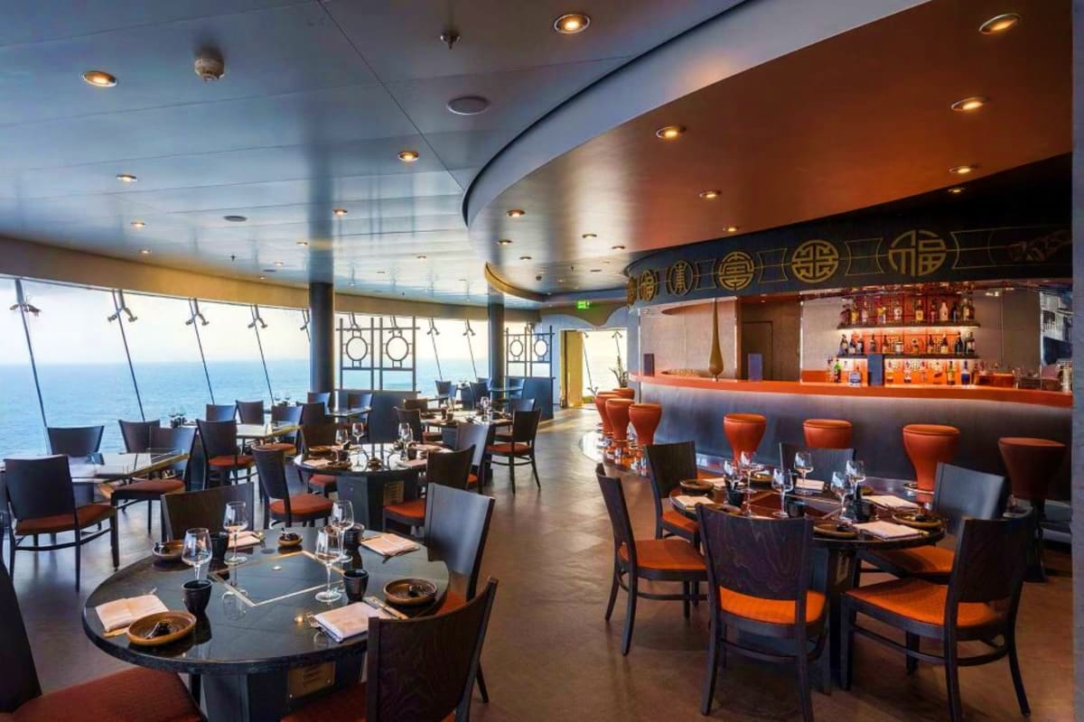 Modern cruise ship restaurant with floor-to-ceiling ocean views, elegant dining tables with orange and black chairs, and full