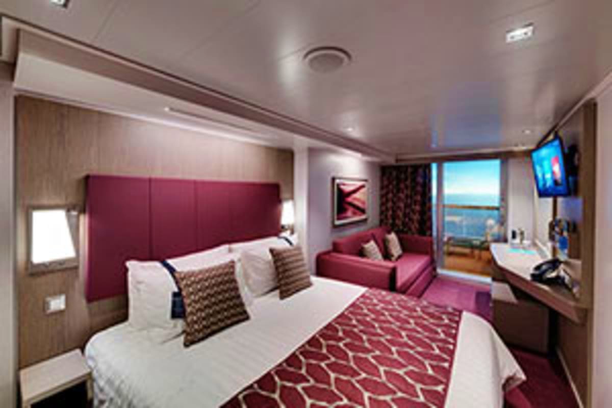 Luxury cruise ship stateroom with burgundy headboard, white bedding, sitting area, flat-screen TV, and ocean view balcony