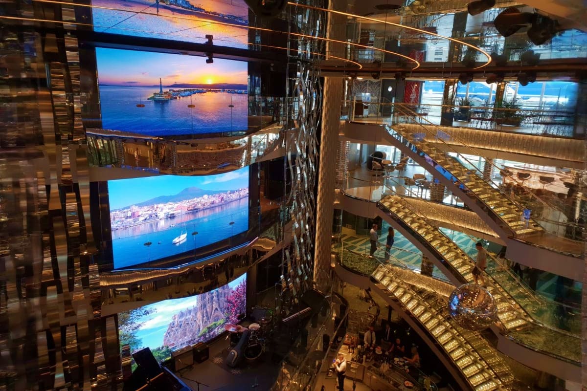 Multi-level atrium with large curved digital screens displaying Mediterranean coastal scenery and sailboats, with passengers 