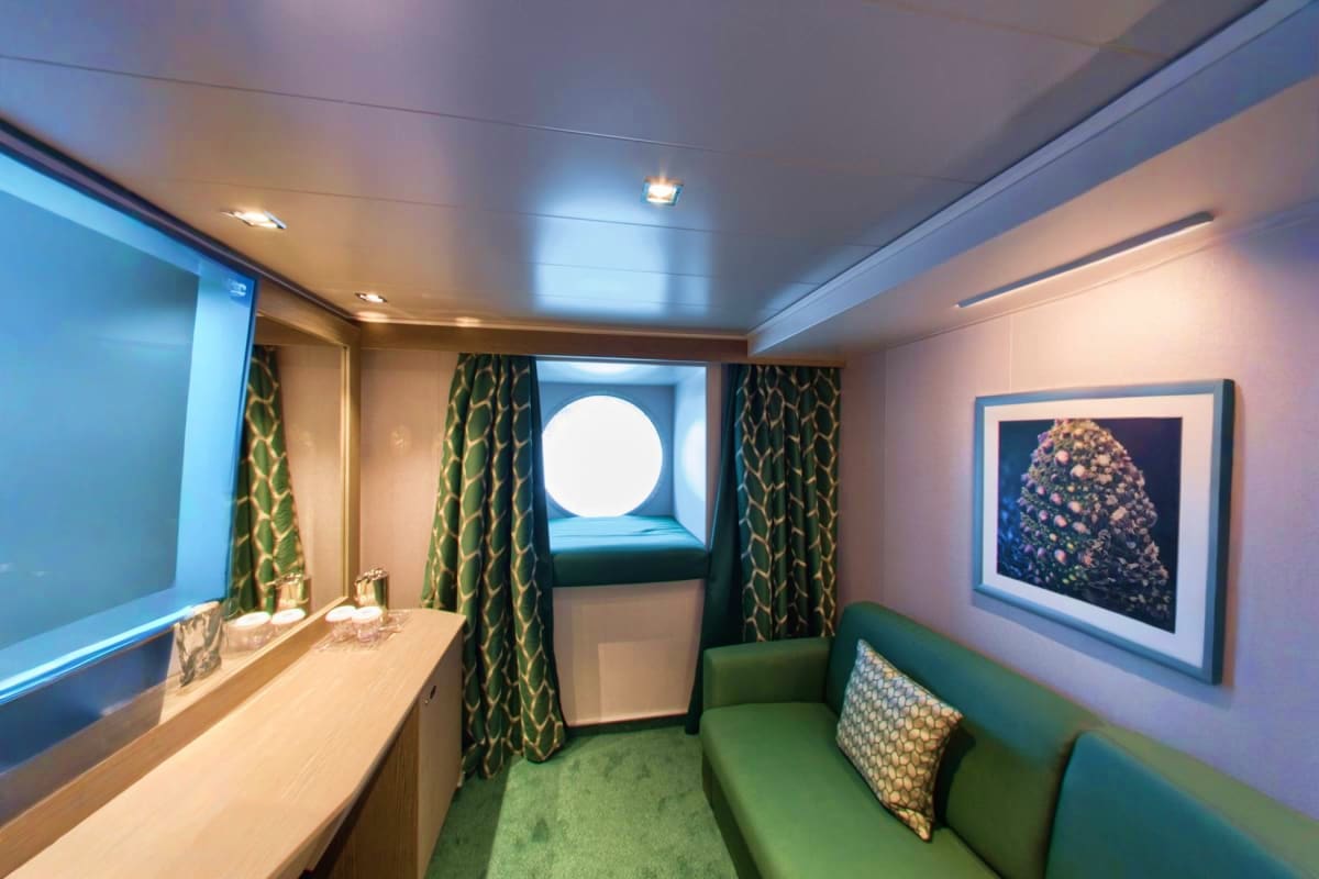 Modern cruise ship cabin with porthole window, green upholstered seating, decorative artwork, and contemporary furnishings