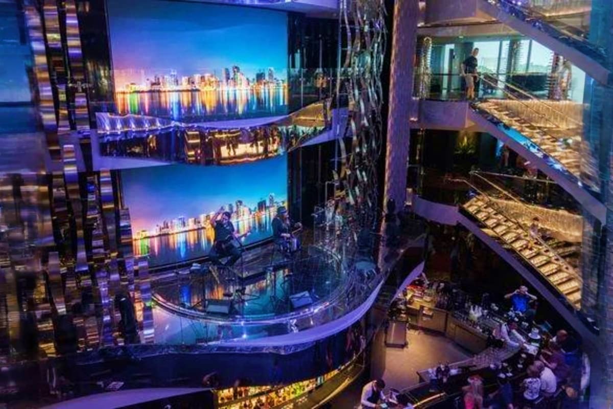 Modern cruise ship interior atrium with curved balconies displaying illuminated cityscape screens showing nighttime harbor wi