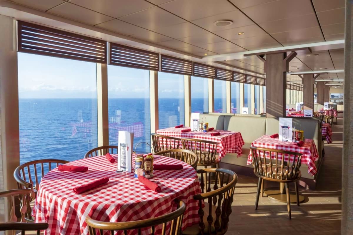 Dining room on cruise ship with red and white checkered tablecloths, ocean views through panoramic windows, and modern interi