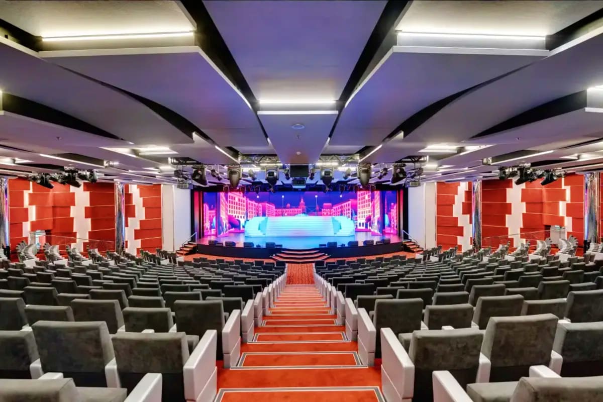 Modern theater auditorium with rows of gray seats, red carpet aisle, illuminated ceiling, and colorful stage set displaying c