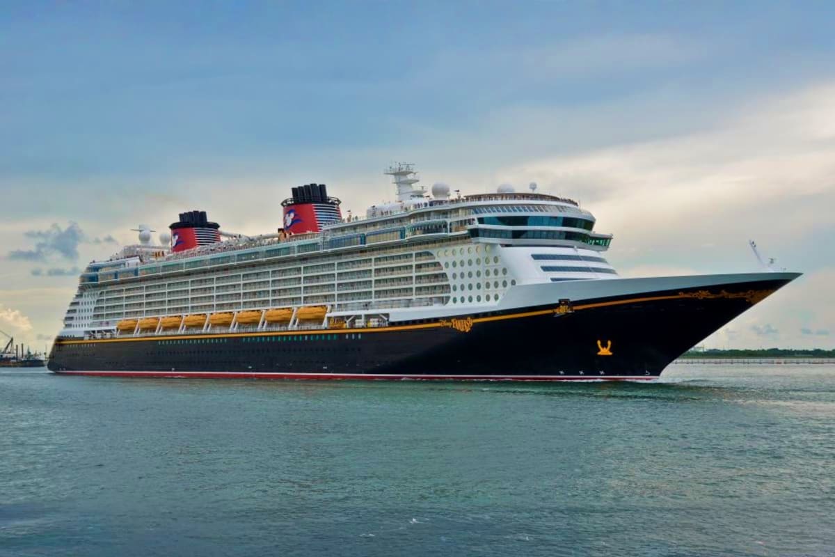 Large Disney cruise ship with black hull and white superstructure anchored in calm turquoise water under cloudy sky
