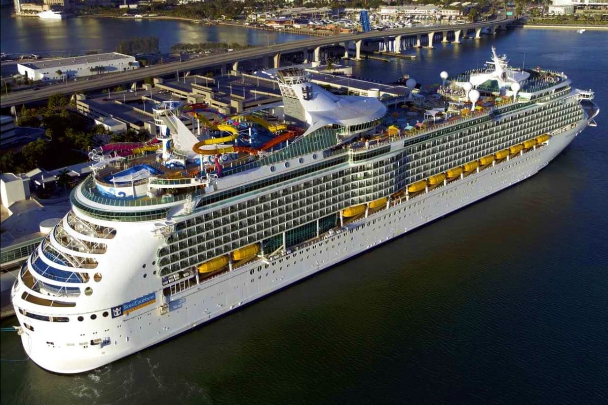 Large cruise ship docked at port with waterslide and recreational facilities visible on upper decks, bridge spanning waterway